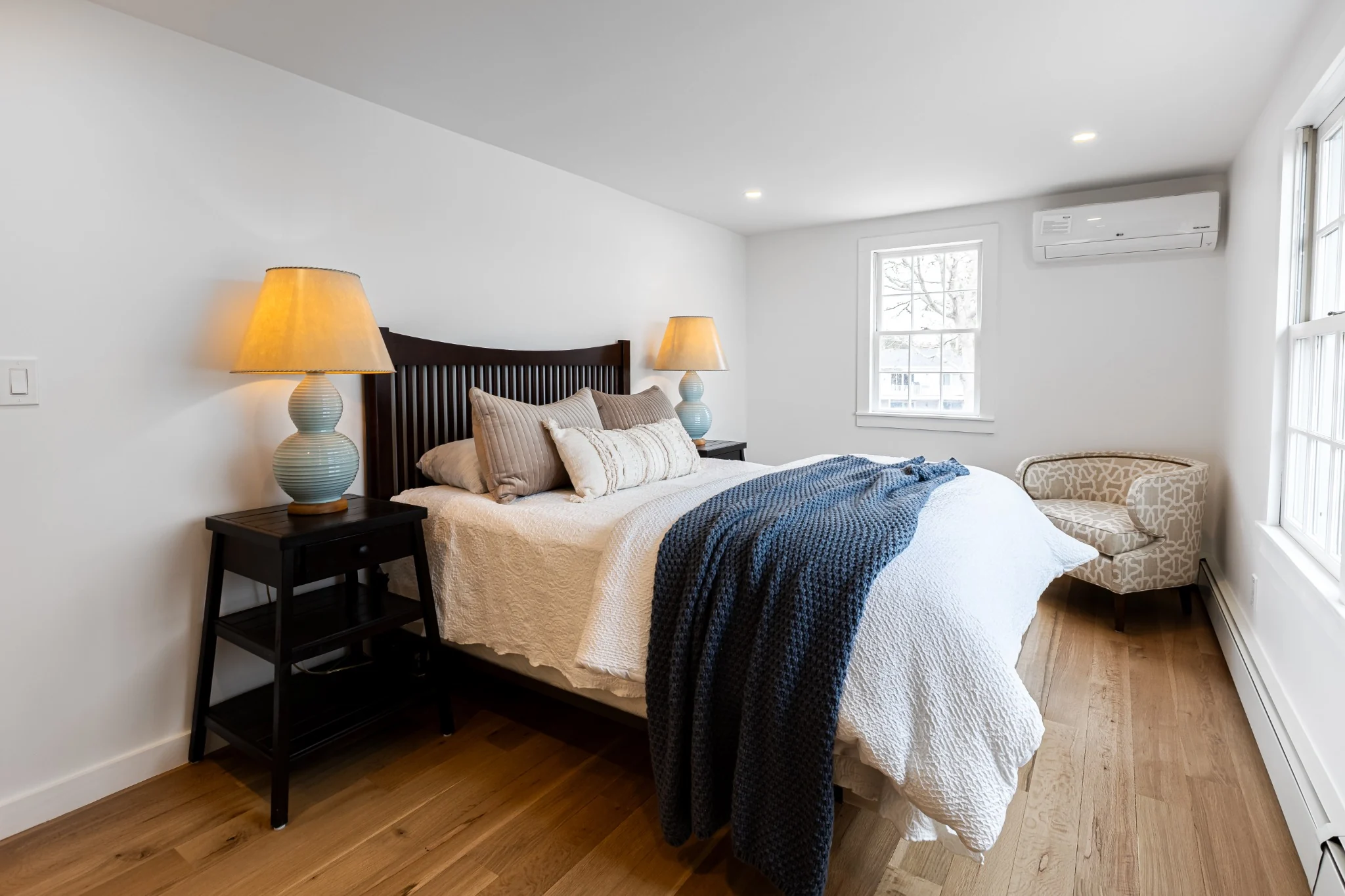 Coastal bedroom with dark wood bed, white bedding, blue throw blanket, and a bright window with water view.