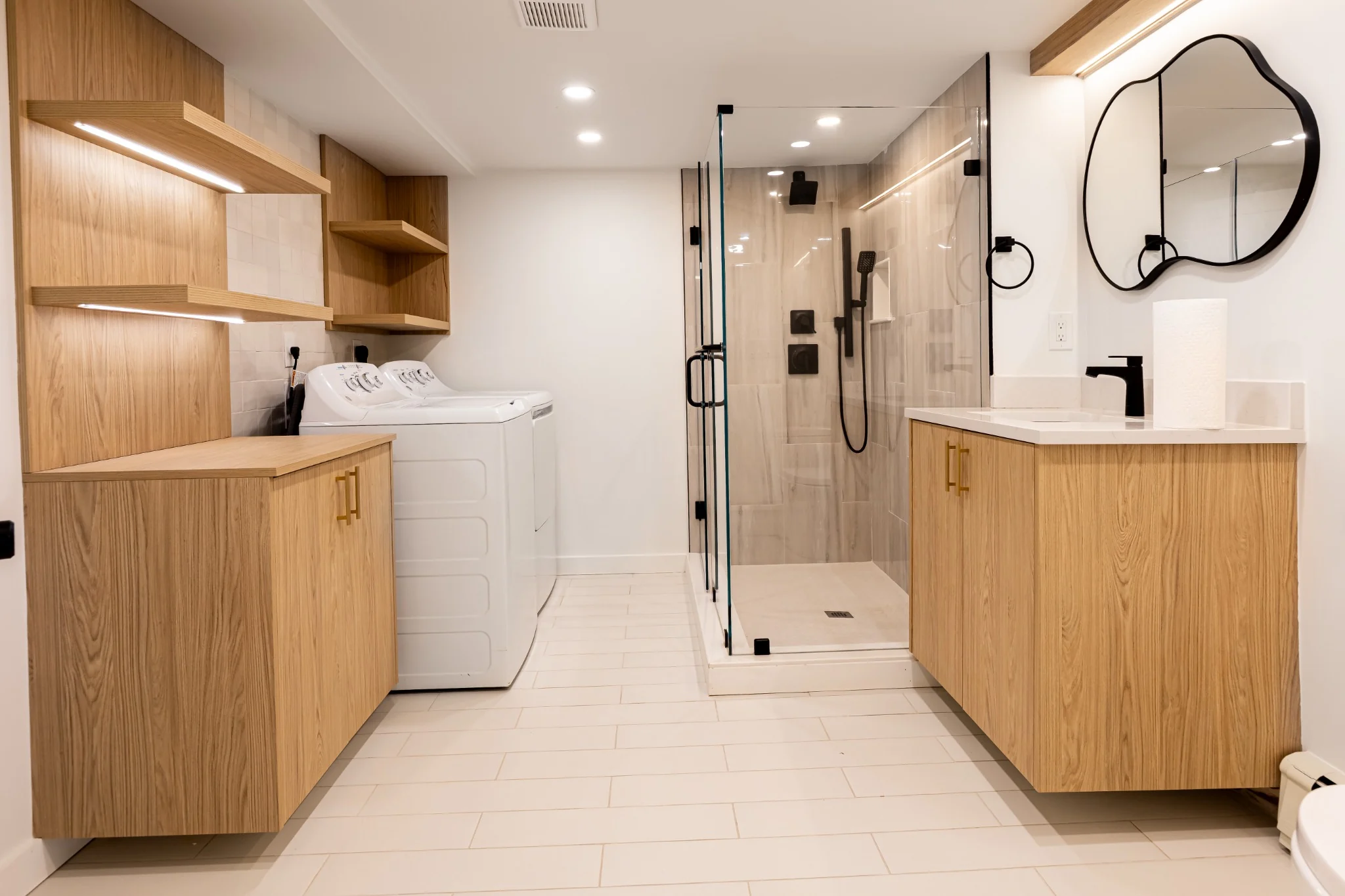Bathroom and laundry room combination with glass shower, clean tile, warm vanity, and modern fixtures.